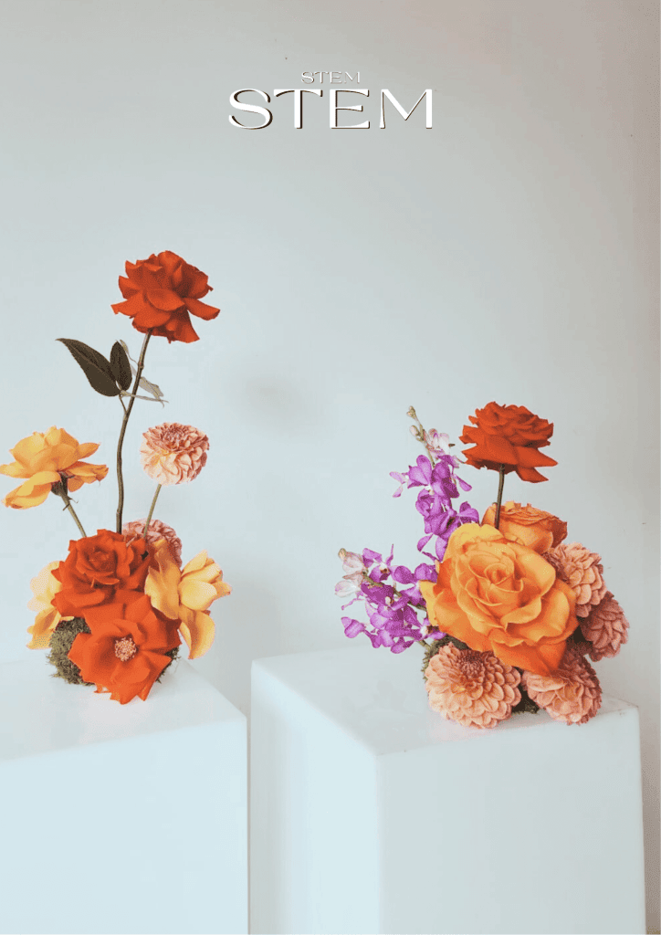 Two floral arrangements with red and orange roses, peach dahlias, yellow garden roses, and purple delphiniums displayed on white plinths, styled by Stem Design Brisbane.