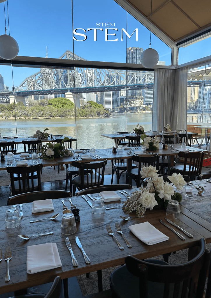 White roses and seasonal greenery arranged in low floral clusters on long timber dining tables at Howard Smith Wharves overlooking the Story Bridge in Brisbane.