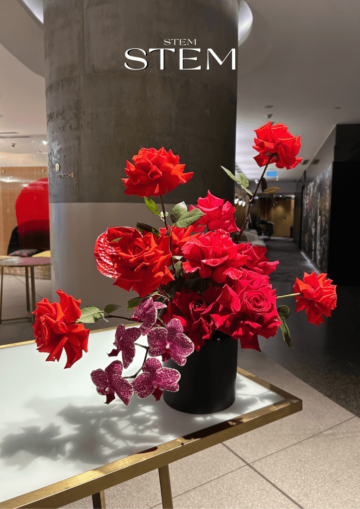 Red and magenta Christmas flower arrangement with reflexed roses and orchids displayed in a black vase.