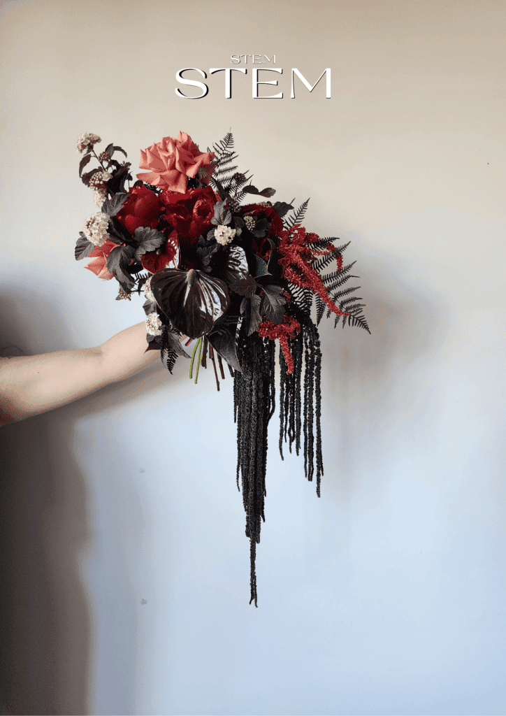 Handheld wedding bouquet with deep red flowers, peach roses, dark foliage and cascading black amaranthus against a neutral background.