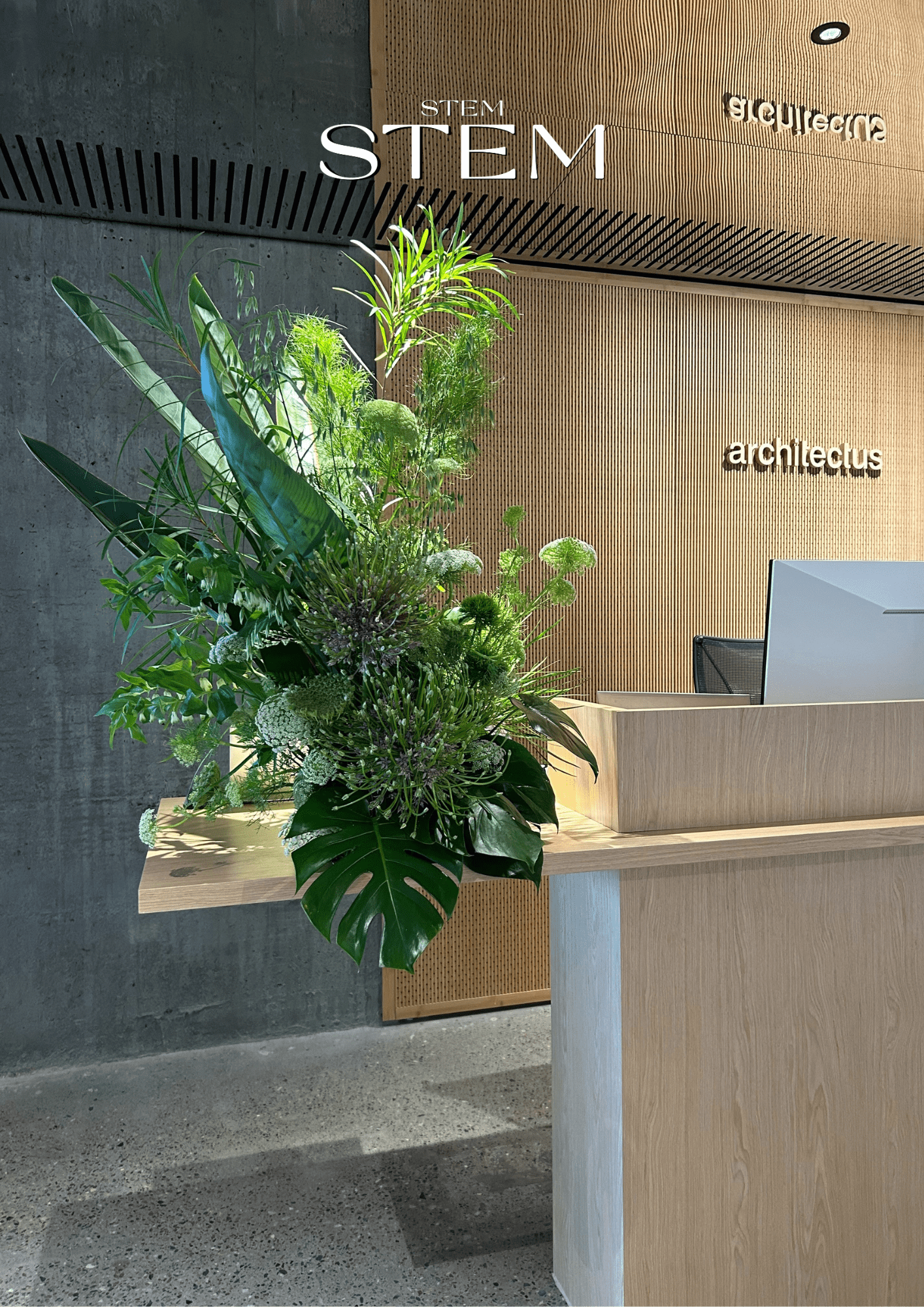 Green corporate flower arrangement with layered tropical leaves and textured seasonal greens on a modern wooden reception desk at Architectus.